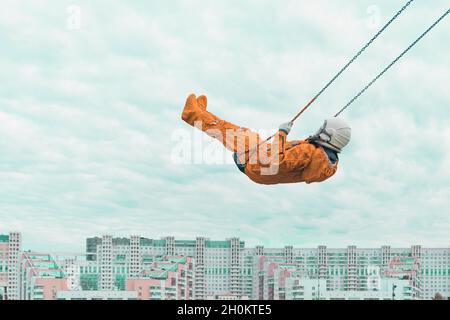 Astronaut wearing orange space suit and space helmet flying on a swing against cloudy sky above the cityscape Stock Photo