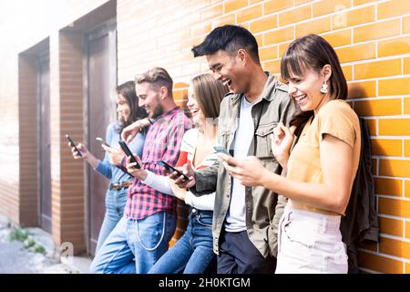 Multicultural friends laughing using smartphone at wall on university college campus - Young people addicted by mobile smart phones - Technology conce Stock Photo