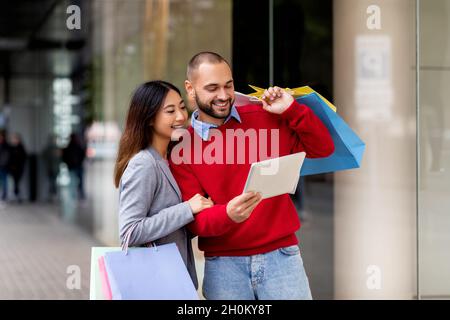 Happy young multiracial couple with gift bags using tablet pc to make online order or check shopping list near city mall Stock Photo