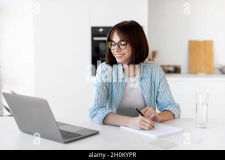 Young freelancer woman working with laptop in kitchen and writing down information while sitting in kitchen Stock Photo