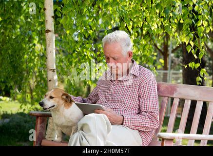 Old man with dog sitting on bench in garden and looking at tablet Stock Photo
