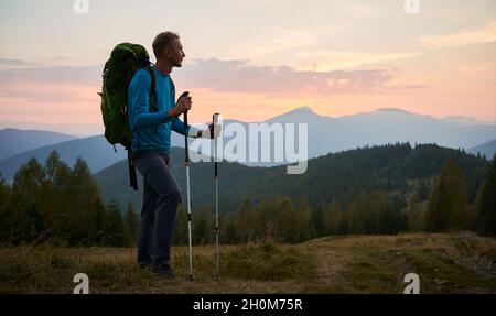 Man traveler against backdrop of mountain hills and pink cloudy evening sky at sunset. Hiker with trekking poles and touristic backpack standing on glade and admiring mountain fairytale landscape. Stock Photo