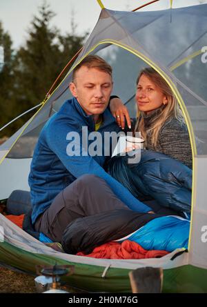 Family couple of mountain campers resting on glade in tent. Husband, lowering his eyes, thinking about next hike. Wife, holding cup, leaning on man's shoulder and smiles enjoying scenery. Stock Photo