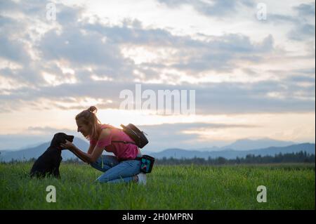 Happy cheerful young woman kneeling down to cuddle her cute black labrador puppy as they are outside on a walk in green meadow. Stock Photo