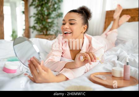 Smiling young african american lady lying on bed, holding and looking at mirror, doing face massage with roller Stock Photo