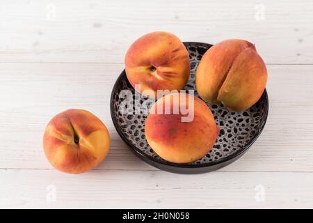 Four fresh ripe juicy peaches are lying in a black clay plate on a white wooden table Stock Photo