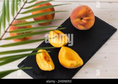 Three fresh ripe orange juicy peaches and their slices are lying on a black rectangular plate and a white wooden table. Top view through palm leaves. Stock Photo