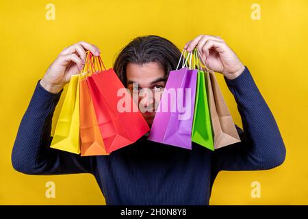 Surprise! Young man on a bright yellow background showing several coloured paper bags with a tempting look. Shopping and gifts concepts. Stock Photo