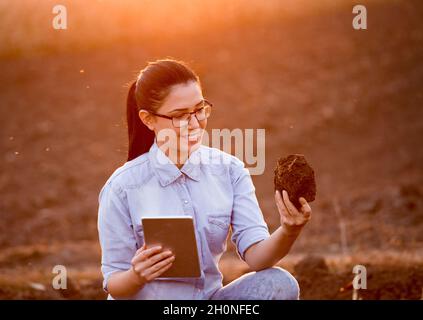 Pretty young engineer woman holding earth clod and tablet and researching soil in field Stock Photo