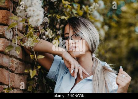 Young Caucasian lady wearing a shirt with accessories posing leaning on a wall with plants Stock Photo