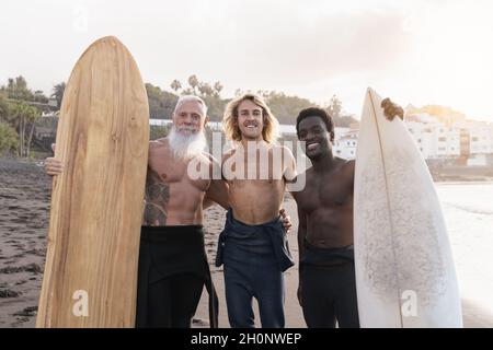 Multiracial surfer friends holding surf boards after extreme water sport session with beach on background - Main focus on senior man face Stock Photo