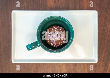 Close up top view of a hot cocoa bomb covered with drizzled brown chocolate and purple and white sprinkles set inside a green coffee mug or cup on rec Stock Photo