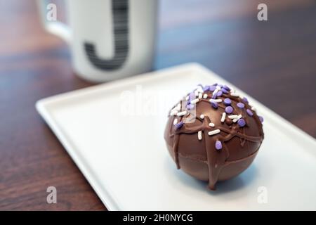 One hot cocoa bomb ball or sphere covered in drizzled brown chocolate and purple and white sprinkles set on a white serving plate with monogram J coff Stock Photo