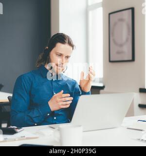 Concentrated young man operator in headset and casual wear talking with client on laptop in office Stock Photo