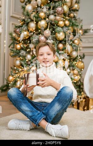 Cute boy is sitting by christmas tree with gifts. Caucasian teenager in warm knitted light sweater sits on the floor of the house with festive new year's decor in the room. Holding gift in his hand Stock Photo