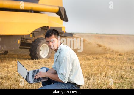 Young engineer with laptop standing in front of combine harvester in field