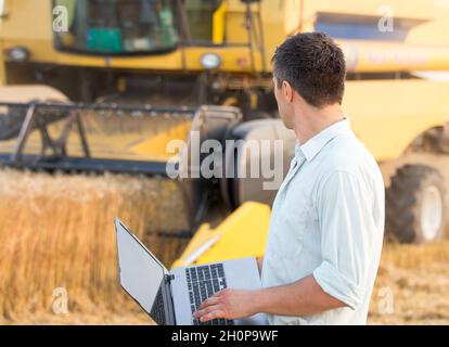 Young engineer with laptop standing in front of combine harvester in field