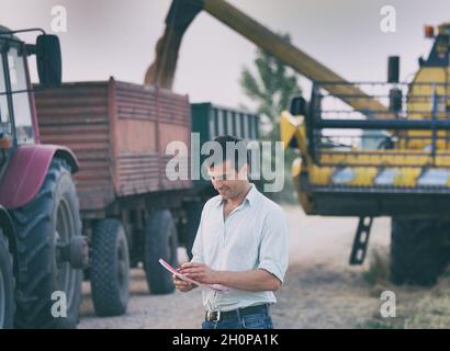 Satisfied young farmer with notebook standing in front of combine harvester while loading trailer with harvested grains in field Stock Photo