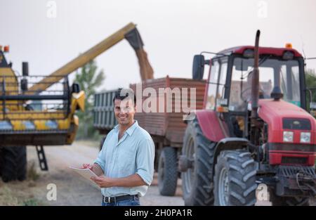 Satisfied young farmer with notebook standing in front of combine harvester while loading trailer with harvested grains in field Stock Photo