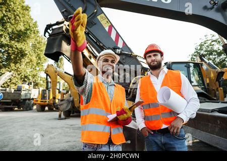 Two men engineers discussing their work standing against construction machines Stock Photo