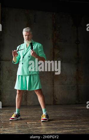 Full length shot of trendy gray haired mature man with beard wearing stylish outfit looking at camera, holding sharp barber scissors and hair comb Stock Photo
