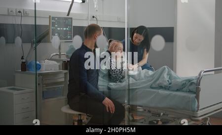 Sick daughter wearing oxygen nasal tube lying in bed after illness surgery waiting for medical examination with parents in hospital ward. Hospitalized child recovering after flu isickness nfection Stock Photo