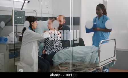 Paediatric doctor examining chest listening heartbeat using medical stethoscope while black woman nurse writing disease treatment working in hospital ward. Kid patient recovery after surgery Stock Photo
