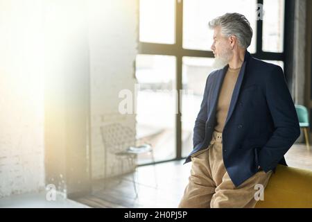 Stylish confident mature man looking aside while posing, leaning on sofa indoors Stock Photo
