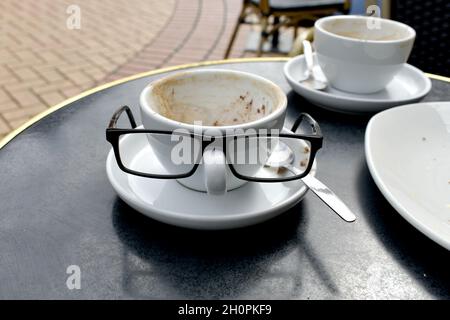 close up of white coffee cups on a table.  His and hers cups with mans glasses perched on handle to look like a funny face outside a café restaurant Stock Photo