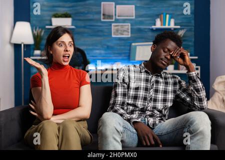 Interracial couple getting into argument sitting at home. Irritated mixed race partners fighting and having relationship dispute. Angry caucasian woman shouting at African american man Stock Photo