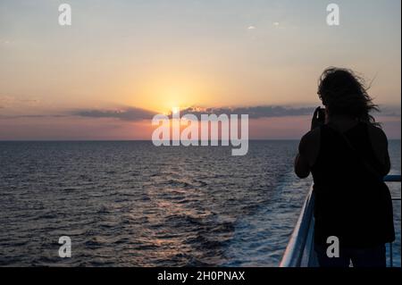 Sunset over the Mediterranean sea viewed from the ferry connecting Toulon and Corsica. Woman on the deck viewed from behind taking a picture of the su Stock Photo