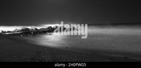Grayscale shot of waves hitting a breakwater in the sea with a sandy beach Stock Photo