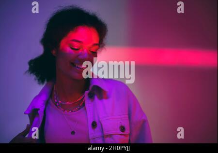 Young happy cheerful african american woman standing under neon glowing light in studio and smiling gently, dressed in denim casual clothes Stock Photo