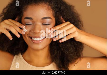 Smiling dark skinned woman with curly afro hair applies face cream on cheeks with both hands, poses over beige studio wall background, uses cosmetic p Stock Photo