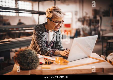 Woman using laptop while standing in carpenter workshop. Female doing male's work concept. Stock Photo