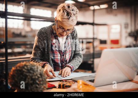 Woman doing paperwork while standing in carpenter workshop. Female doing male's work concept. Stock Photo