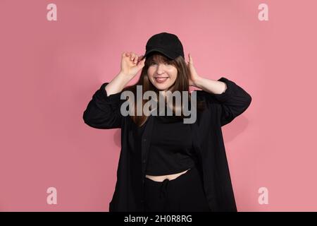 portrait of dancing young woman on pink background in studio with baseball cap and black sportswear looks into frame, fervently and cheerfully. Stock Photo
