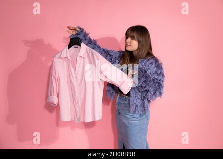 young woman fashion stylist holds hanger with shirt on pink background in studio. Basic wardrobe concept. Blogger teaches classes on creating stylish Stock Photo