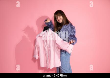 young woman fashion stylist holds hanger with shirt on pink background in studio. Basic wardrobe concept. Blogger teaches classes on creating stylish Stock Photo