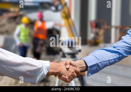 Two business people shaking hands in front of leveling instrument on construction site with workers in background Stock Photo