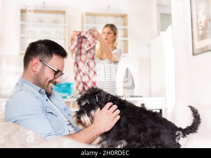 Young handsome man sitting on sofa and cuddling dog while wife ironing and doing chores in background Stock Photo