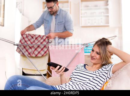 Beautiful young woman resting on sofa and watching tv while man doing laundry in background. Husband doing chores concept Stock Photo