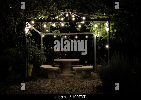 A lit arbor in a green garden in the middle of the night. With a table and chairs made of stone. Stock Photo