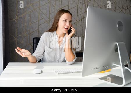 Young freelancer woman is talking on smartphone working with laptop from home, female employee has pleasant phone conversation working on the distance, connect with customers by mobile phone Stock Photo