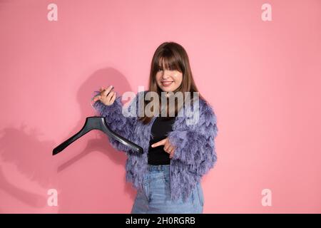 Blogger teaches classes on creating stylish image. A young pretty woman holds empty clothes hanger on pink background in studio. Stock Photo