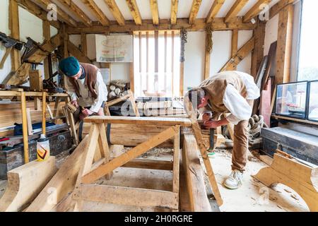 Two men, medieval reenactors, working at both ends of a large piece of oak wood in a re-created carpentry workshop. Both chiselling out square holes. Stock Photo