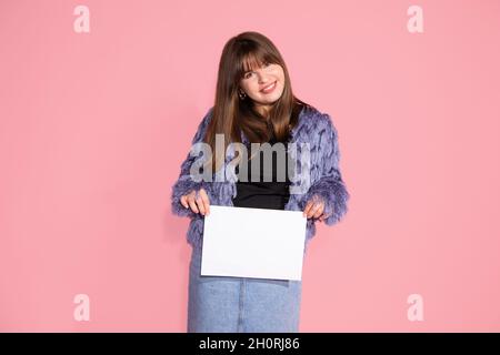 Beautiful young dark-haired woman with bang on pink background in studio holding blank sheet of paper with copy space. Smiling at camera Stock Photo