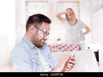 Smiling handsome man typing on mobile phone on sofa while angry housewife ironing in background at home Stock Photo
