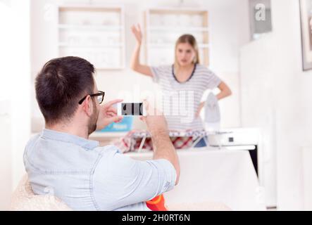 Handsome young man having fun capturing his angry girlfriend ironing and doing chores Stock Photo