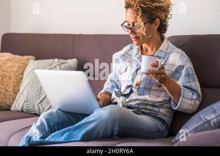 Confident happy adult young woman use laptop computer sitting on the sofa at home in relax leisure activity drinking coffee or tea and smiling - moder Stock Photo
